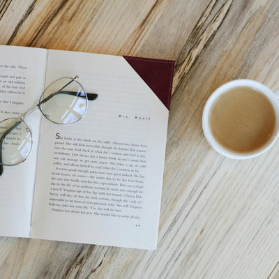 Open book with glasses on a wooden surface next to a cup of coffee